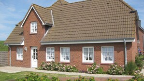 A two-story brick house with a sloped roof and a front garden with flowering plants. - Guesthouse Am Badedeich, vacation apartment Sylt in Dagebüll for up to 4 people (Dagebüll)