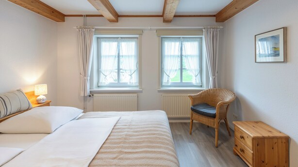 A bedroom with a bed, two windows with curtains, a chair, a wooden nightstand with a lamp, and a framed picture on the wall. - Guesthouse Am Badedeich, vacation apartment Sylt in Dagebüll for up to 4 people (Dagebüll)