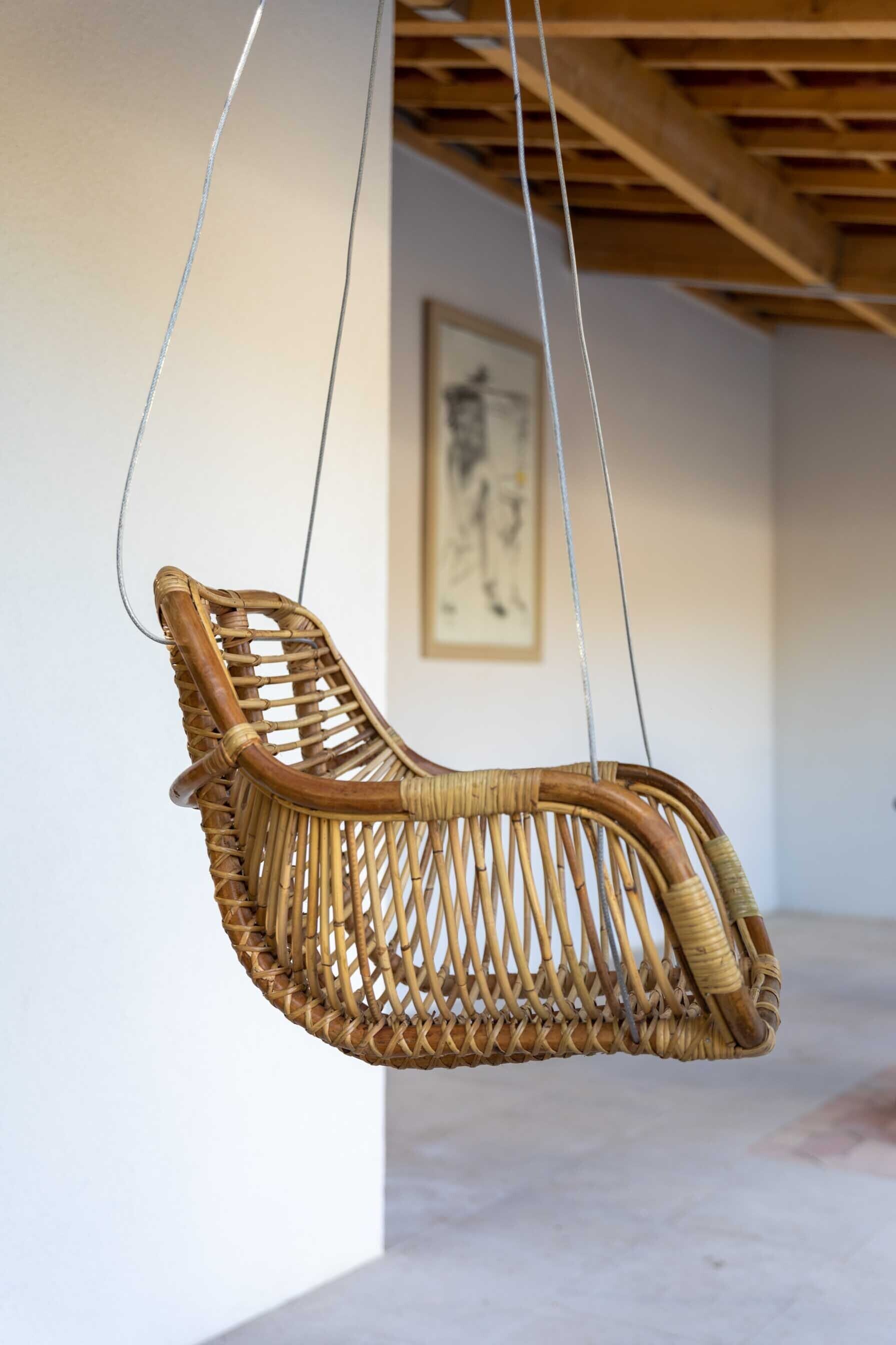 Ein Korbstuhl, der an Seilen aufgehängt ist, in einem Raum mit Holzboden und einem gerahmten Kunstwerk an der Wand.