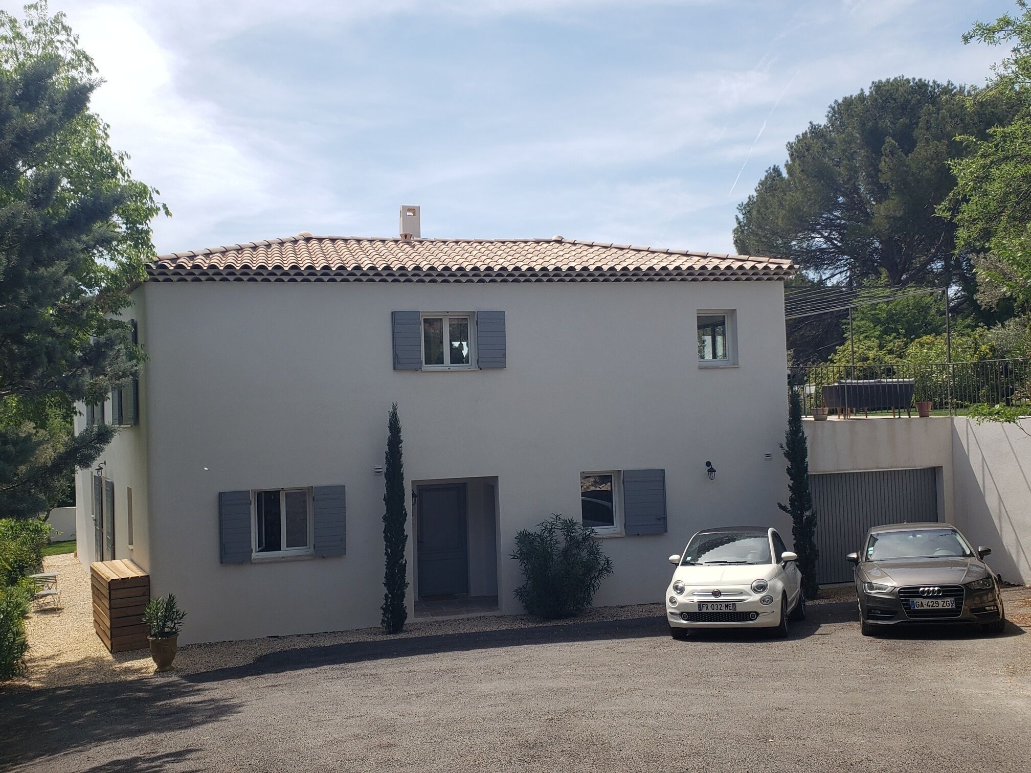 A two-story house with a tiled roof, two cars parked in front, and a gravel driveway.