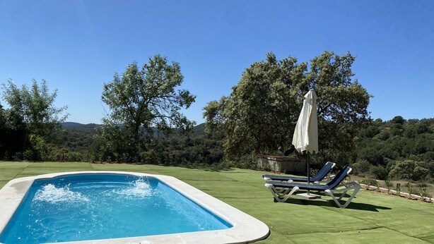 A rectangular swimming pool with a white edge, surrounded by a green lawn. There are two lounge chairs with white umbrellas and a small table. The background features a hilly landscape with trees.