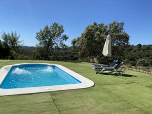 Ein rechteckiger Pool mit weißem Rand, umgeben von einer grünen Rasenfläche. Zwei Liegestühle mit weißen Sonnenschirmen und ein kleiner Tisch stehen bereit. Im Hintergrund ist eine hügelige Landschaft mit Bäumen zu sehen.
