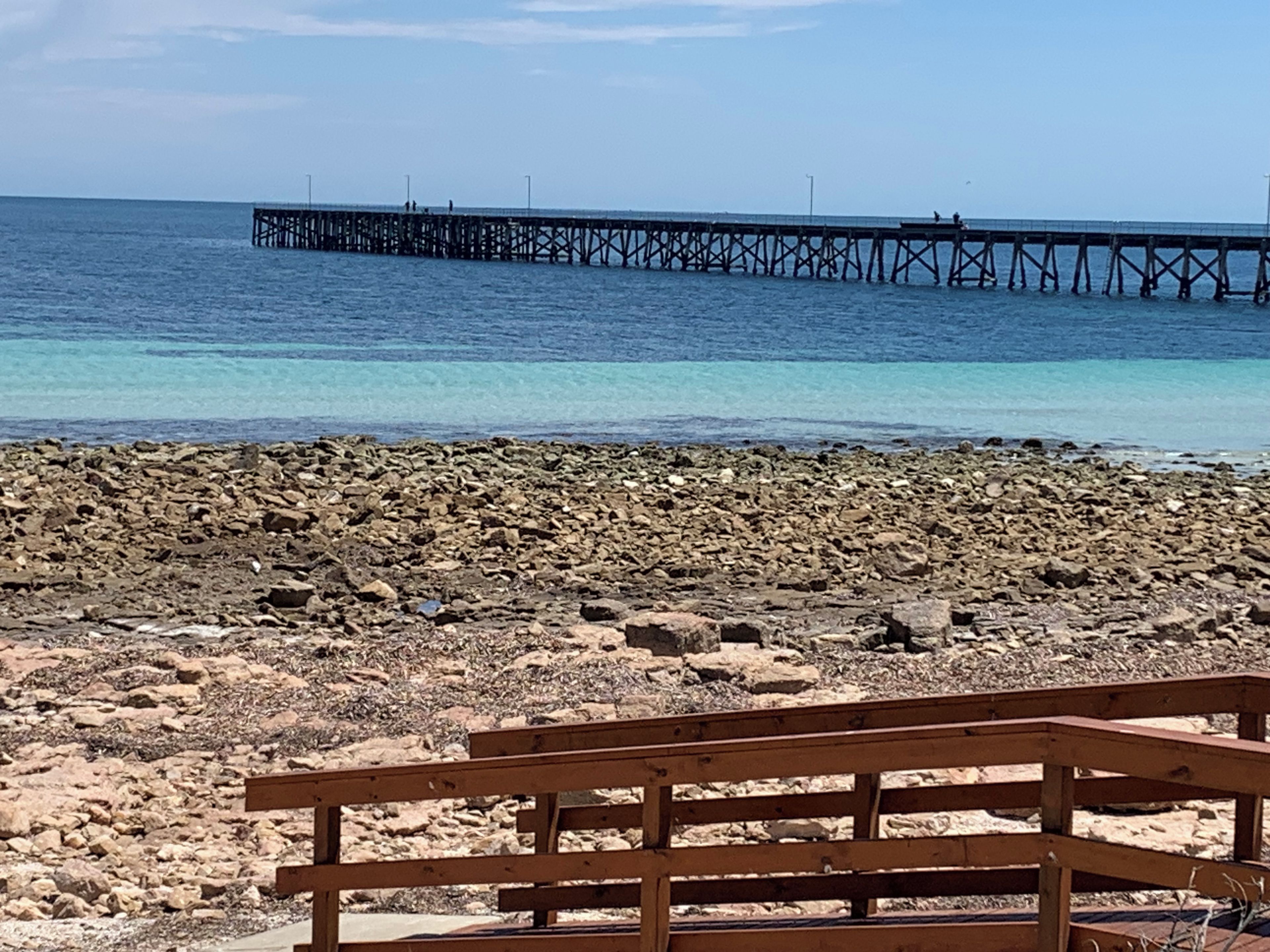 Une plage rocheuse avec un banc en bois au premier plan et une jetée qui s’avance dans l’eau à l’arrière-plan.