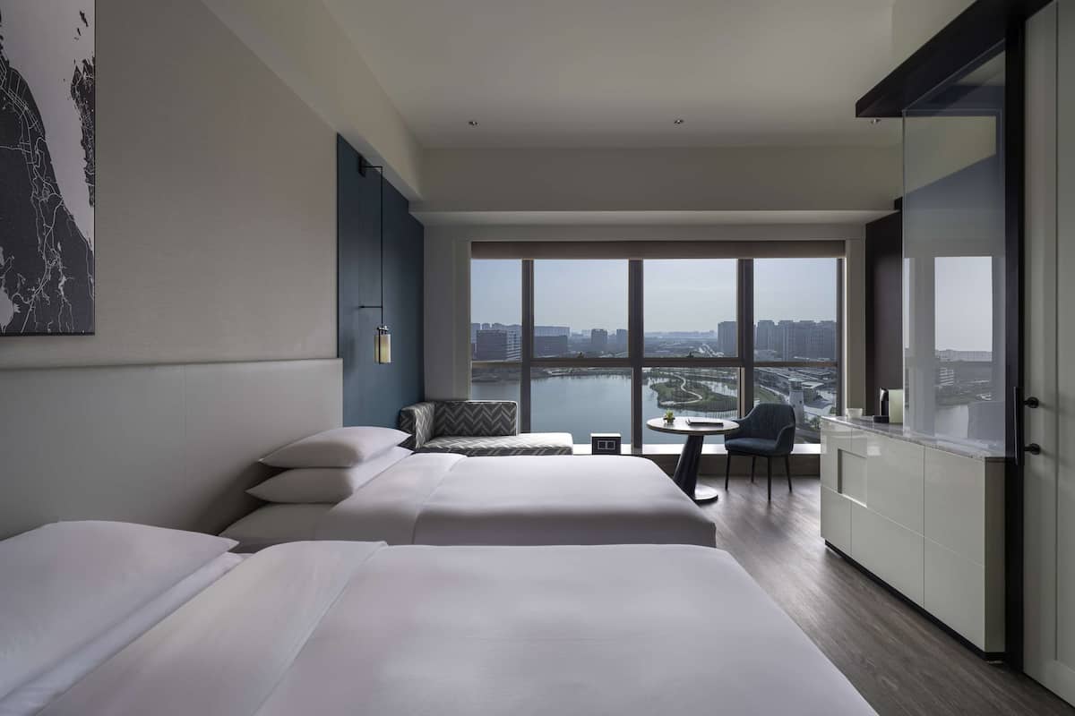 Une chambre d’hôtel moderne dotée d’une grande fenêtre donnant sur la ville, d’un lit, d’un coin salon et d’une petite table.