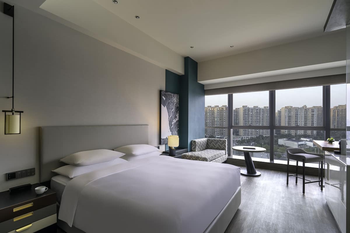 Une chambre d’hôtel moderne dotée d’un grand lit, d’un coin salon avec une table et offrant une vue sur le paysage urbain grâce à de grandes fenêtres.