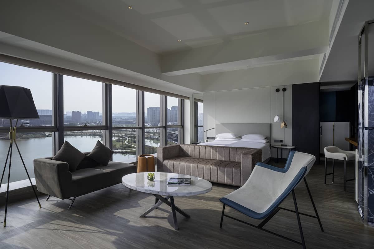 Une chambre d’hôtel moderne avec une grande fenêtre, un canapé, un fauteuil, une petite table et un lit.