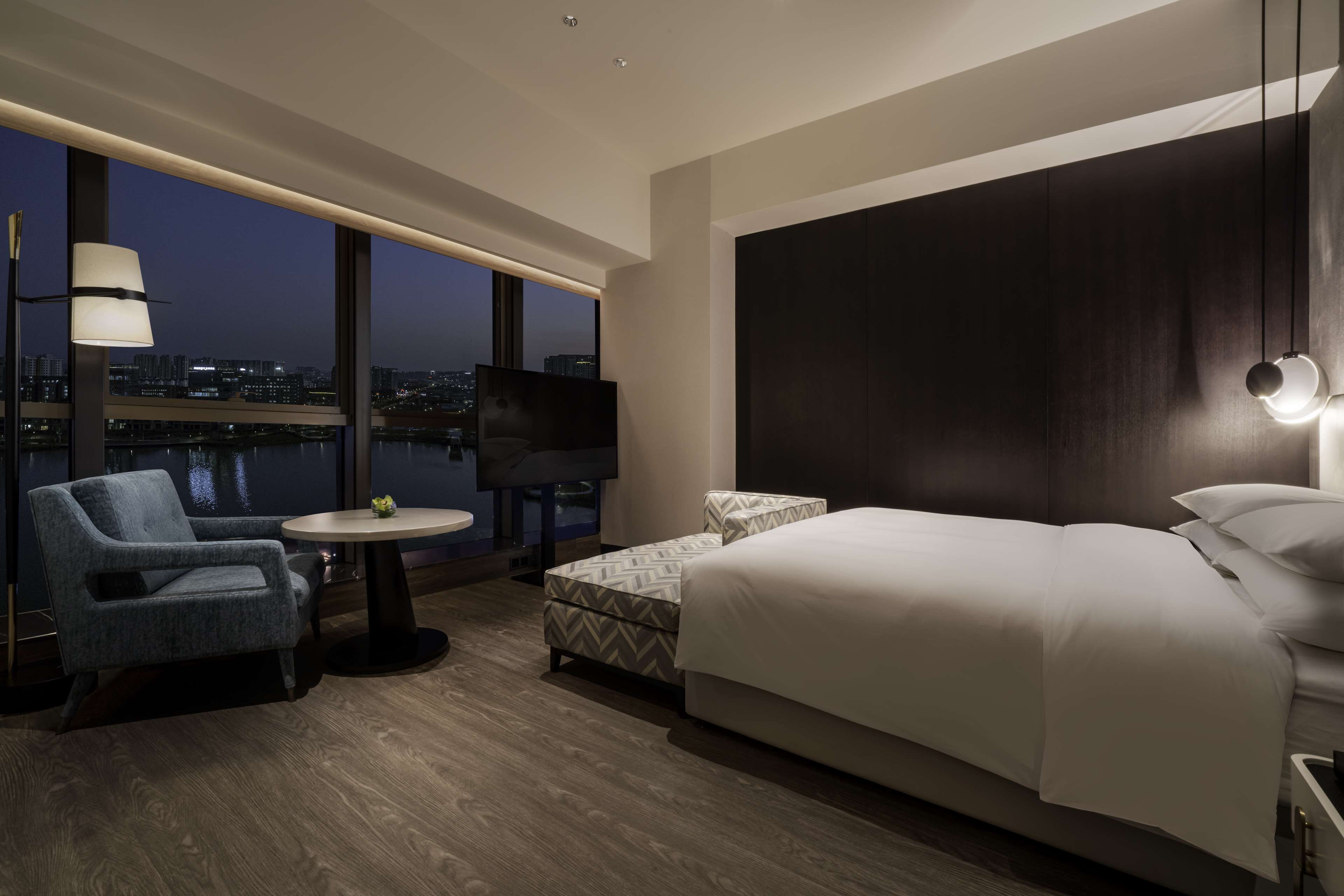 Habitación de hotel moderna con una cama grande, zona de estar con una silla y una mesa, televisor de pantalla plana y vistas a la ciudad a través de amplias ventanas.