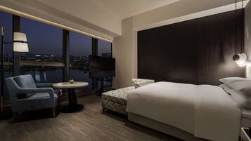 Habitación de hotel moderna con una cama grande, zona de estar con una silla y una mesa, televisor de pantalla plana y vistas a la ciudad a través de amplias ventanas.