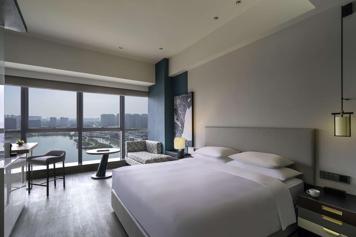 Une chambre d’hôtel moderne dotée d’un grand lit, d’un bureau avec une chaise, d’une petite table et offrant une vue sur le paysage urbain grâce à de grandes fenêtres.