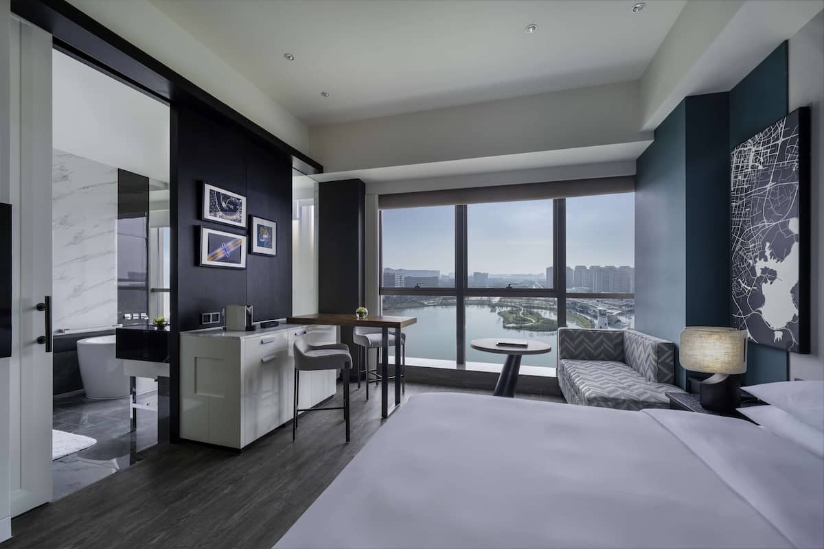 Une chambre d’hôtel moderne dotée d’un grand lit, d’un bureau avec une chaise, d’un canapé et offrant une vue sur le paysage urbain grâce à de grandes fenêtres.