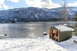Een blokhut aan een meer, omringd door sneeuw en met bergen op de achtergrond.