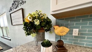 Un taulell de cuina amb una planta en test, un gallinet decoratiu i una citació emmarcada a la paret.