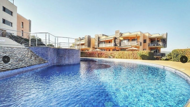 A swimming pool with a stone border and a tiled edge, surrounded by a building with balconies and a clear sky. - Private property in front of Boadella beach (Lloret de Mar)