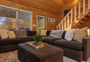 A cozy cabin interior with a large brown sectional sofa, a wooden coffee table with a plant, and a staircase with a 'HEY Y'ALL' sign.