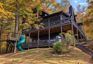 A multi-level wooden cabin with a deck and a slide, surrounded by autumn foliage. - Enchanted Hideaway: TreeHouse • Slide • HotTub • FirePit • Fun Games • Secluded (Blue Ridge)