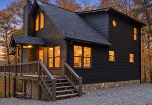 A two-story house with a steeply pitched roof, a front porch with a staircase, and a stone foundation.