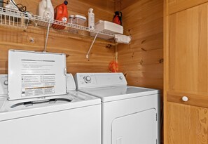 A laundry area with a white washing machine and dryer, a shelf with cleaning supplies, and a wooden wall.