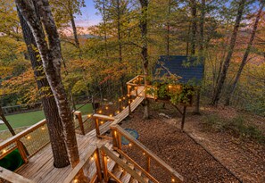 A wooden deck with a rope bridge, surrounded by trees and a blue building.