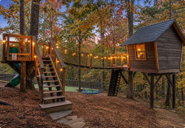 A wooden cabin with a staircase leading to a platform, surrounded by trees and string lights.