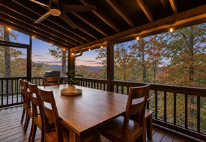 Un área de comedor al aire libre con una mesa y sillas de madera, un ventilador de techo y vistas a follaje otoñal.
