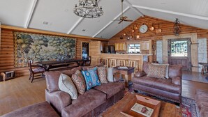 A spacious living room with a high ceiling, wooden walls, and a large painting. There is a dining area with a wooden table and chairs, and a kitchen in the background. - Guadalupe Bluff Log Cabin 4 Bedroom Home by RedAwning (Kerrville)