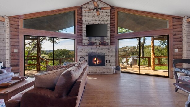A spacious living area with a fireplace, leather sofa, and large windows offering a view of the outdoors. - Guadalupe Bluff Log Cabin 4 Bedroom Home by RedAwning (Kerrville)