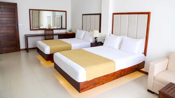 Habitación de hotel con dos camas, cabecero de madera, mesita de noche con lámpara, espejo y puerta de madera.