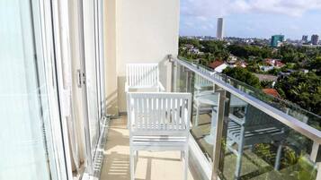 A balcony with two white chairs, a glass railing, and a view of a cityscape with buildings and greenery.