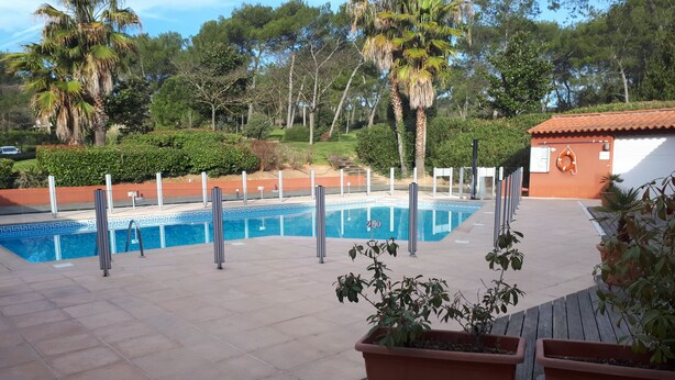 A swimming pool with a tiled deck, surrounded by palm trees and a red-roofed building. - Appartement piscine (Mouans-Sartoux)