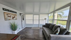 Una habitación amplia con un gran ventanal, un sofá gris, suelo de madera y una puerta con un pequeño ventanuco.