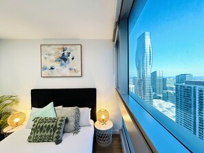 Une chambre avec un grand lit, une vue sur la ville, un tableau abstrait encadré et une table de chevet avec une lampe.
