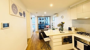 A modern kitchen with white cabinets, a gas stove, and a built-in oven. There is a small island with a sink and a chair. The kitchen has wooden flooring and a view of the balcony through the window.