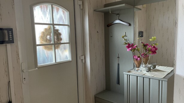 Une salle de bain avec une porte blanche, une fenêtre à vitre dépolie, un meuble-lavabo avec un miroir, une serviette suspendue et un vase avec des fleurs.