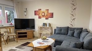 Un salon avec un canapé gris, un meuble TV en bois et un téléviseur à écran plat. On y trouve une petite table en bois recouverte d’une nappe en dentelle et un bol décoratif. Une fenêtre avec des rideaux est visible, ainsi qu’une étagère murale où sont disposés divers objets.