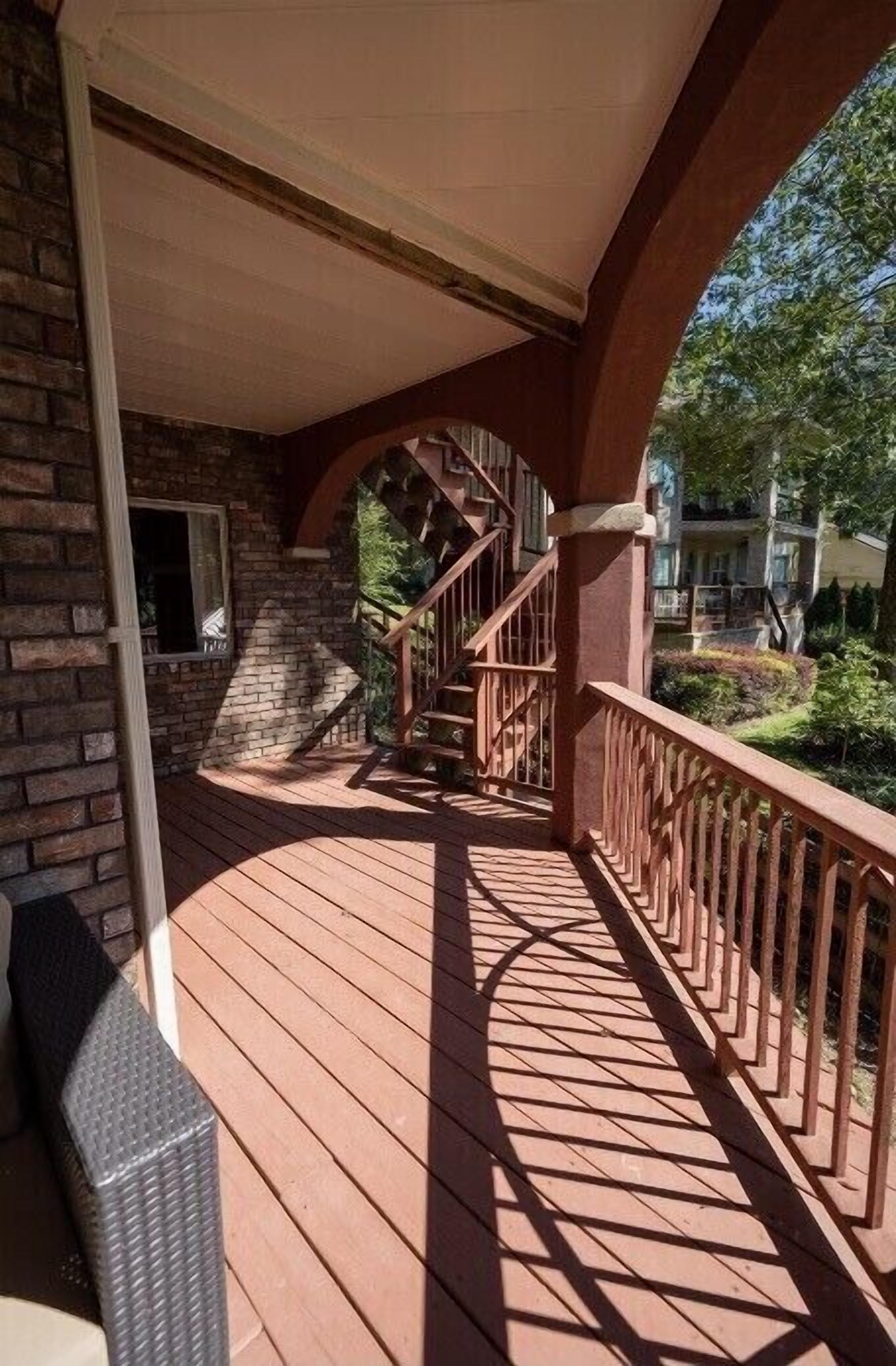 A wooden deck with a staircase leading to a covered area, surrounded by brick walls and greenery.