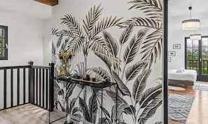 A hallway with a wall mural of tropical leaves, a black-framed console table with decorative items, and a staircase with a wooden railing.