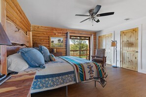 A bedroom with a wooden ceiling, a bed with a quilt, a chair, and a window with a view of trees.