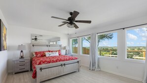 Une chambre à coucher comprenant un lit, un ventilateur de plafond, une table de chevet, une lampe et de grandes fenêtres donnant sur le paysage.