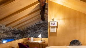 A cozy attic bedroom with a sloped ceiling, a stone wall, a wooden desk, a purple chair, and a bed with a decorative headboard.