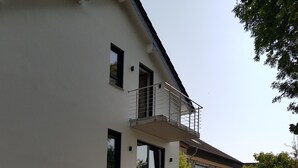 Un edificio moderno de dos pisos con un balcón, un camino de entrada pavimentado y vegetación circundante.