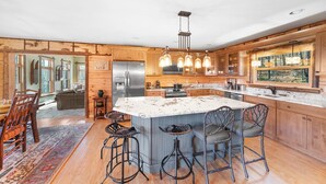 A spacious kitchen with wooden cabinets, a large island with bar stools, a stainless steel refrigerator, and a hanging light fixture. - Private Lincoln Log Luxury Lodge in the Adirondacks  (Johnsburg)