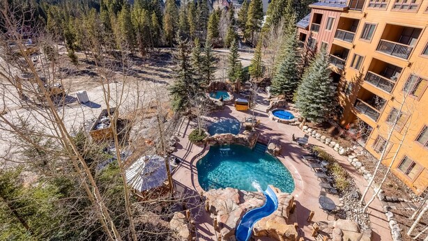 A resort with a pool area, including a hot tub and a slide, surrounded by trees and mountains in the background. - Spacious Top-Floor 3-bed Springs Condo: 5 minute walk to gondola and restaurants (Keystone)