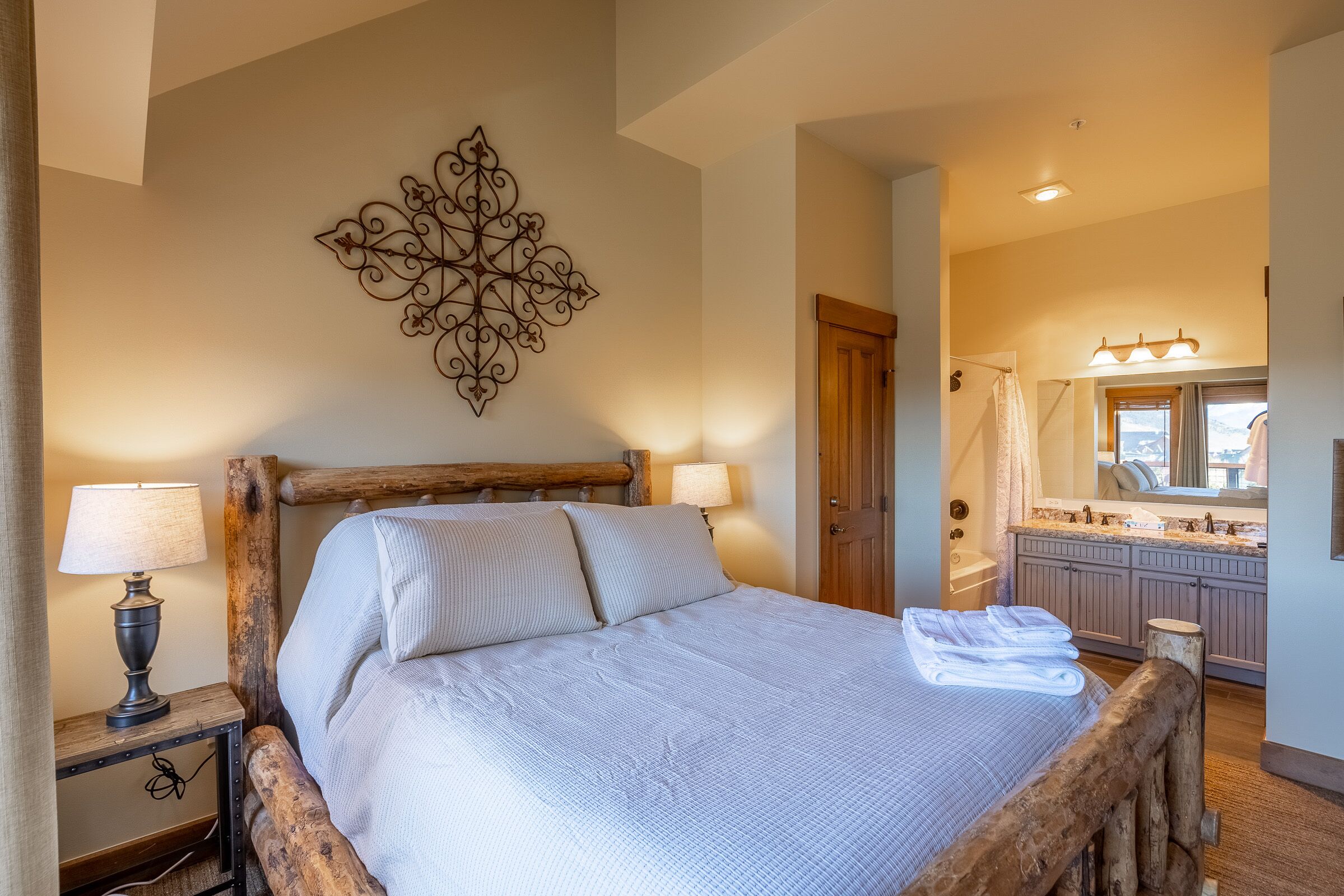 Un lit avec une tête de lit en bois, un élément décoratif fixé au mur et une salle de bain avec un meuble-lavabo et un miroir.