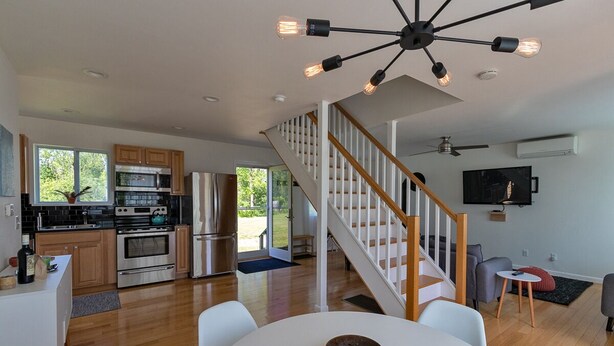 A modern kitchen with stainless steel appliances, a dining area with a white table, and a staircase leading to a living area with a TV. - Peaceful River Retreat with Fireplace & Panoramic Views (Cementon)
