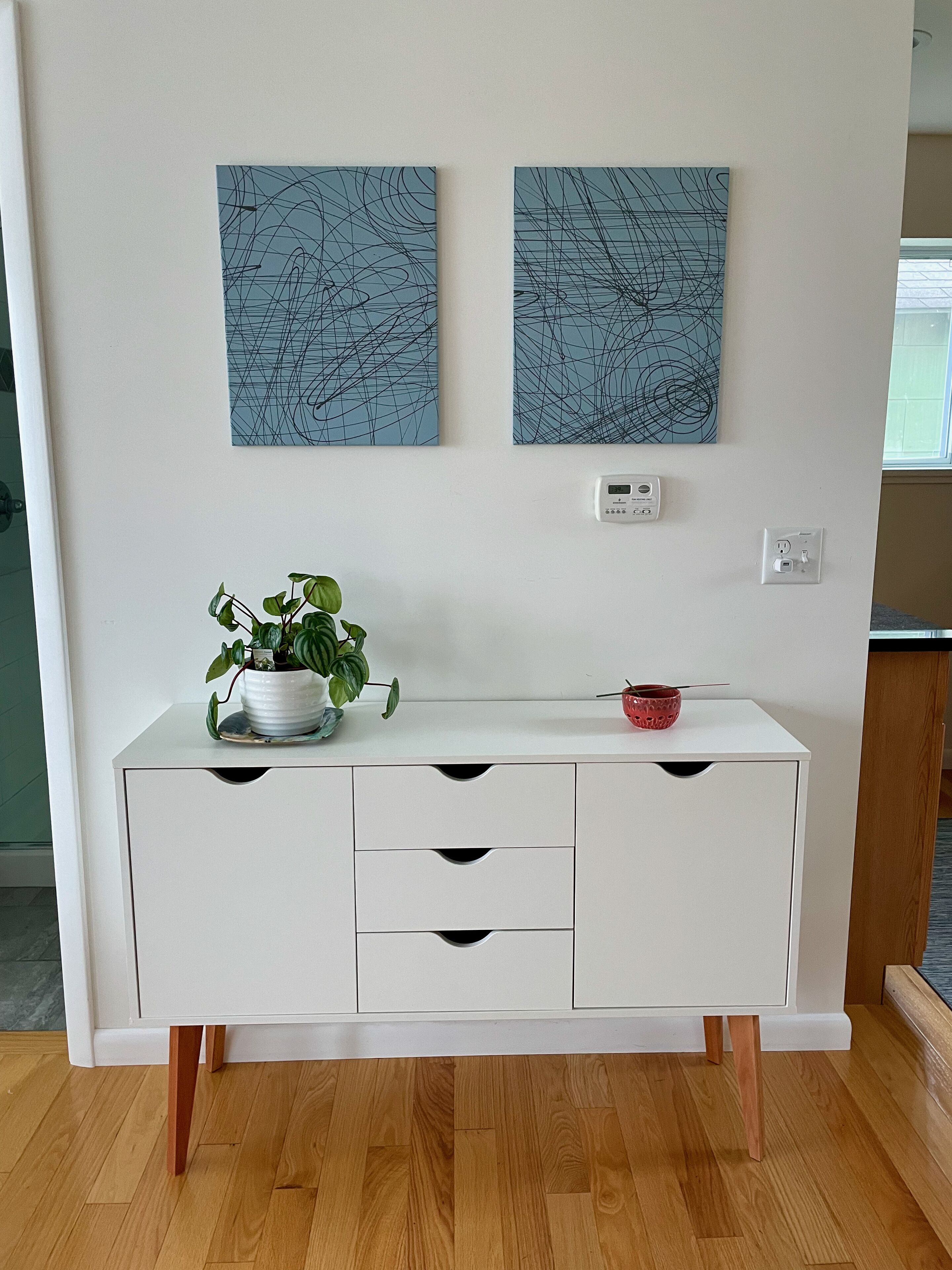 Een witte kast met drie lades, een plant in een pot en een rode kom bovenop. Daarboven hangen twee abstracte schilderijen.