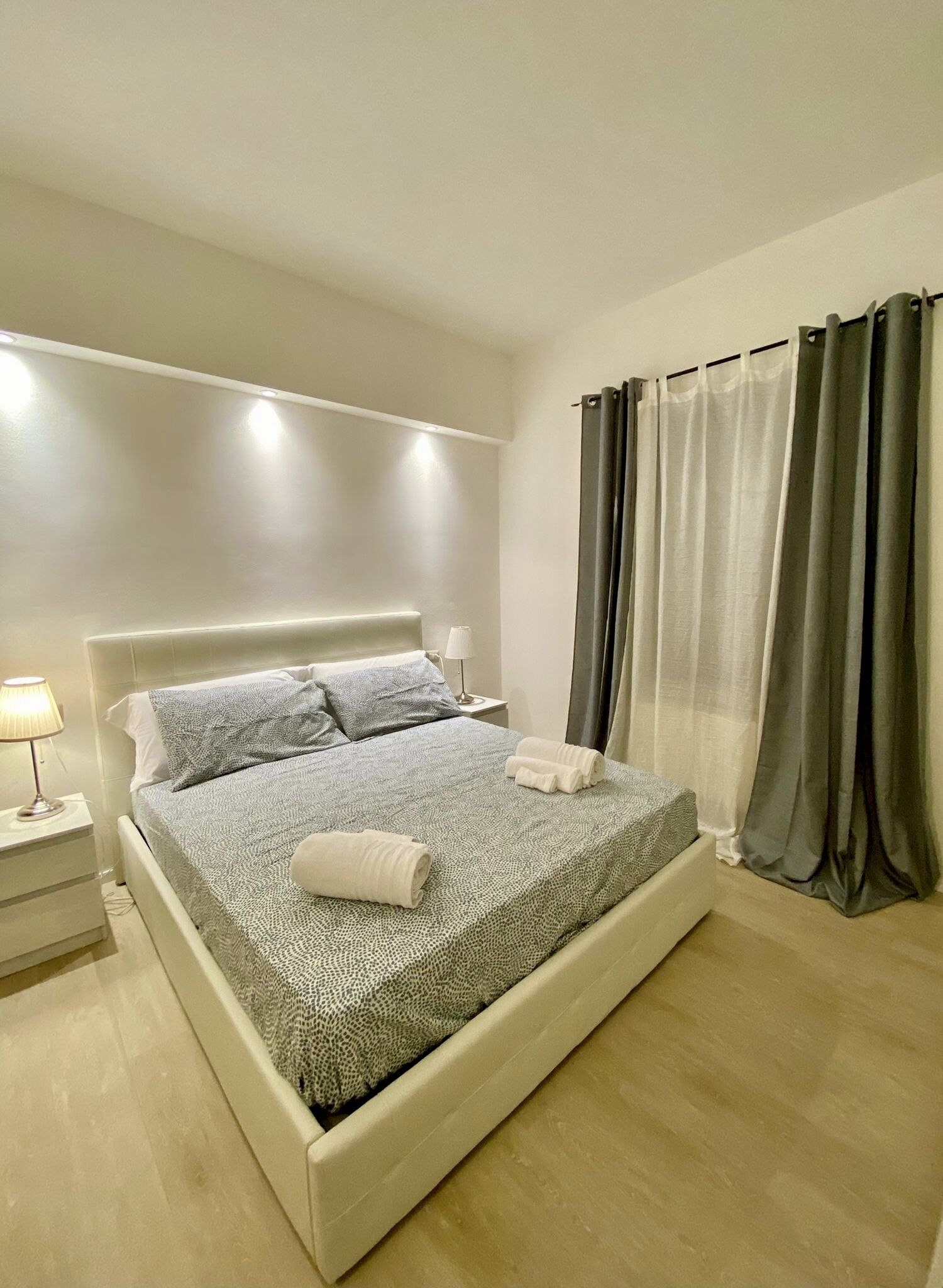 Ein modernes Schlafzimmer mit einem großen Bett, einem Nachttisch mit Lampe und einem Fenster mit Vorhängen.