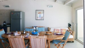 Uma sala de jantar com uma mesa e cadeiras de madeira, uma cozinha com geladeira e uma janela grande.