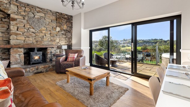 A modern living room with a stone fireplace, leather furniture, and a large sliding glass door offering a view of the countryside. - Cartref - Beautiful Cottage Hot Tub Countryside Views (Saundersfoot)