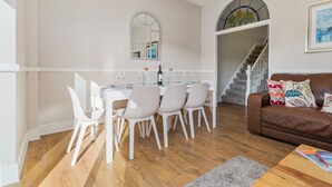 A dining area with a white table and chairs, a wine bottle, and a mirror above the table. There is a staircase leading to an upper level, and a brown leather sofa with patterned cushions in the foreground. - Cartref - Beautiful Cottage Hot Tub Countryside Views (Saundersfoot)