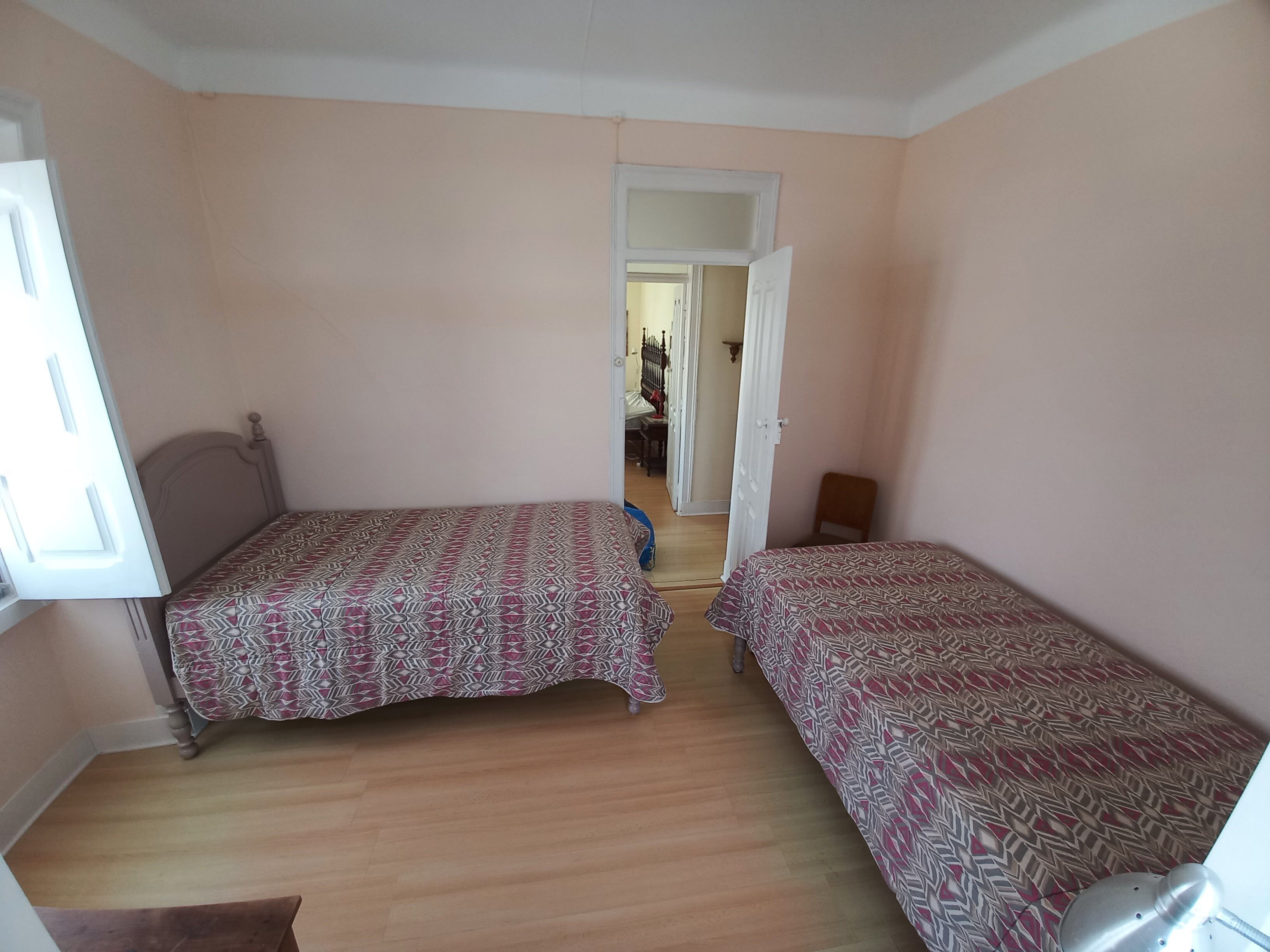 A room with two beds, wooden flooring, and a door leading to another room.